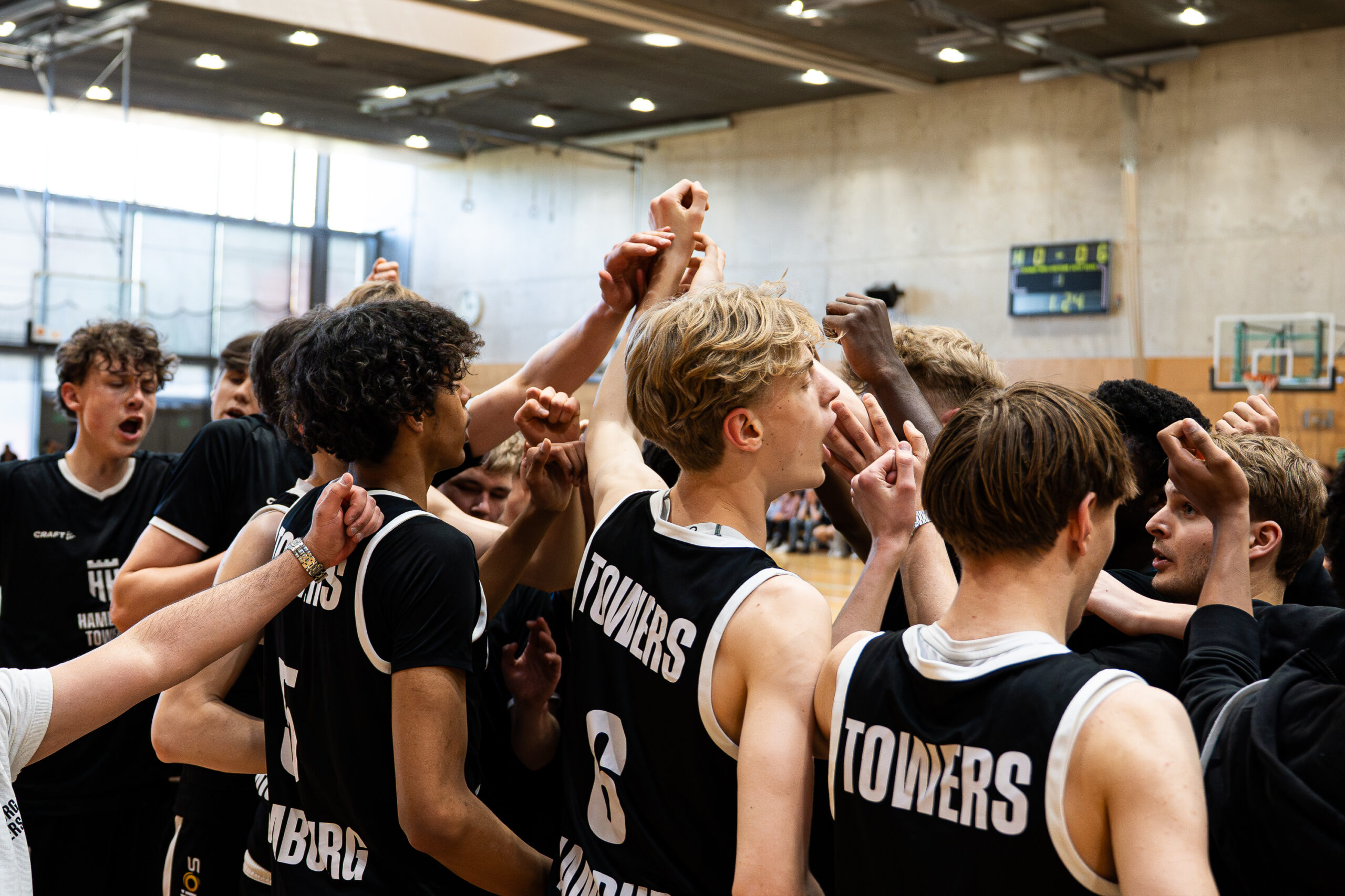 Basketballspieler der Hamburg Towers in schwarzen Trikots mit hochgereckten Armen in einer Teamrunde in der Halle
