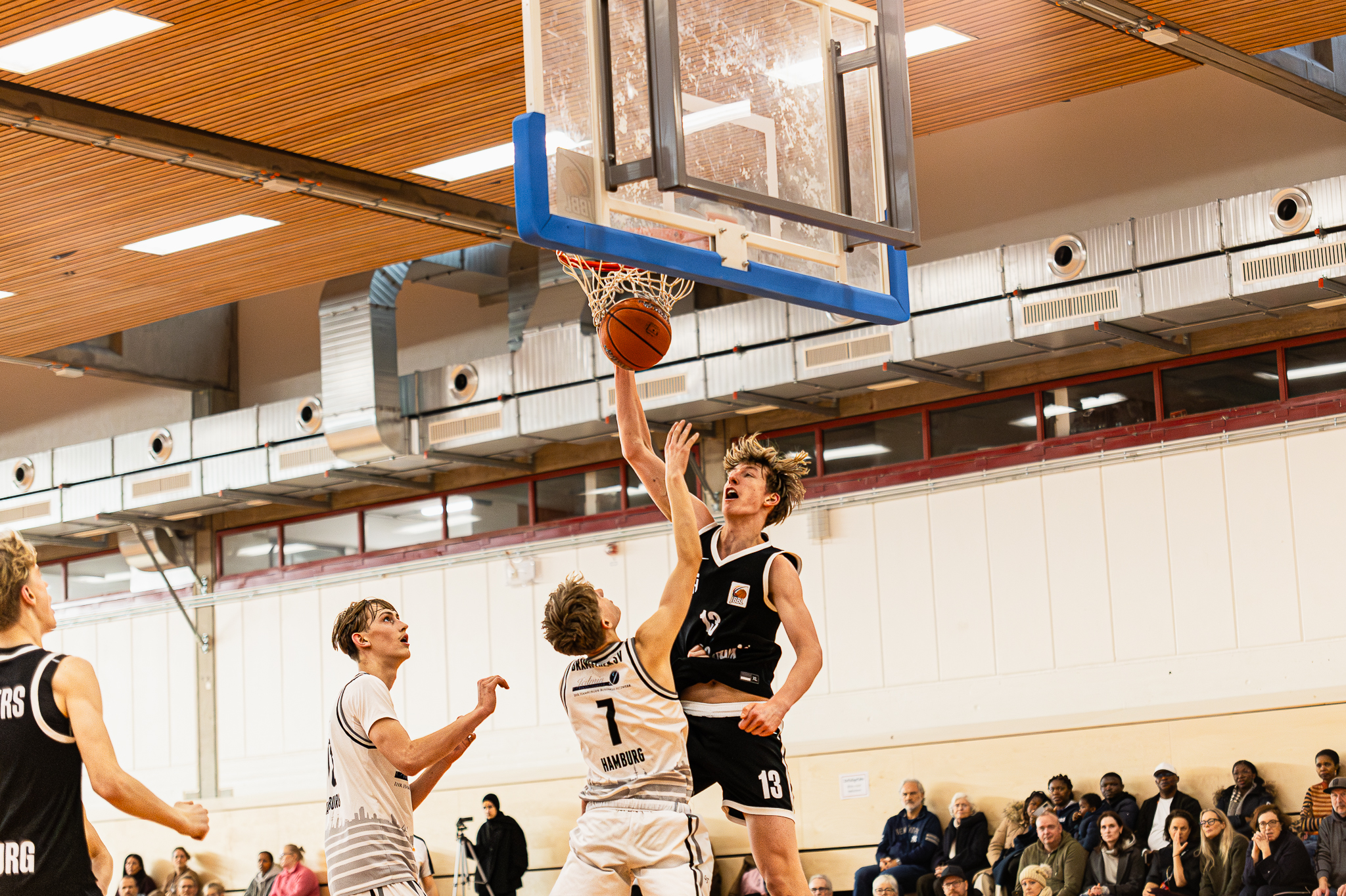 Mehrere Basketballspieler in Aktion unter einem Korb in einer Halle mit Zuschauern auf der Tribüne