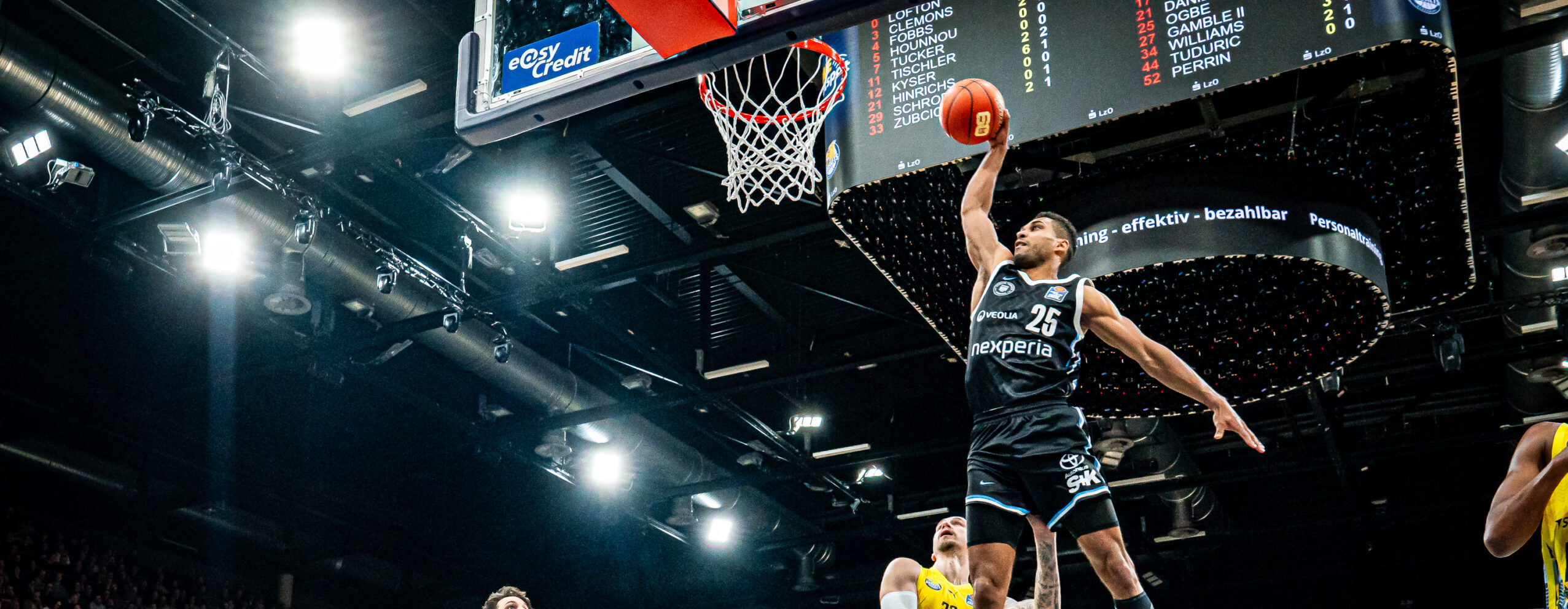 Basketballspieler in schwarzer Sportkleidung springen zum Korb in einer Halle mit Publikum und Werbebannern