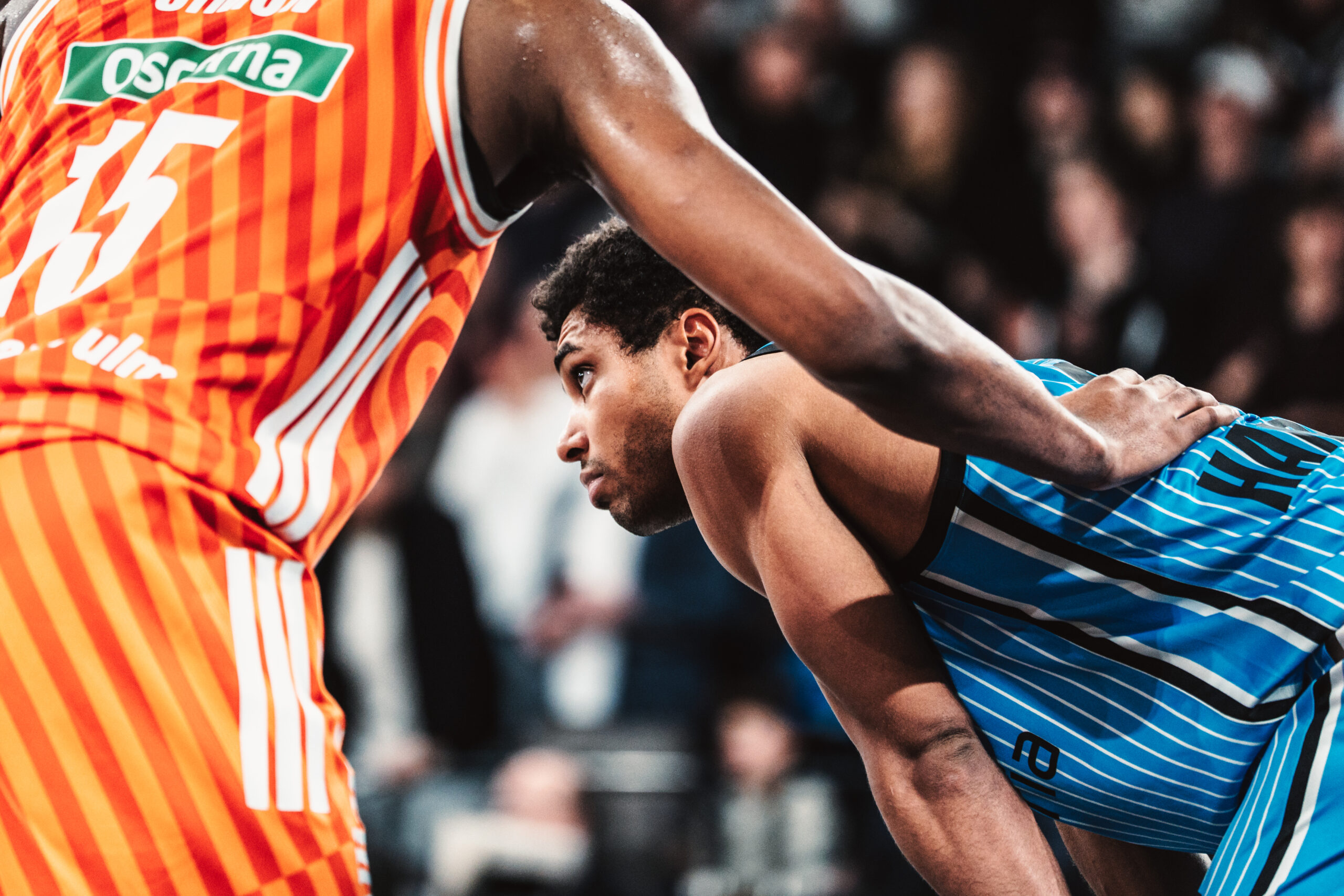 Zwei Basketballspieler in Aktion, einer in orange-weiß gestreiftem Trikot mit der Nummer 15, der andere in blau-schwarzem Trikot mit der Nummer 25, der sich vorbeugt