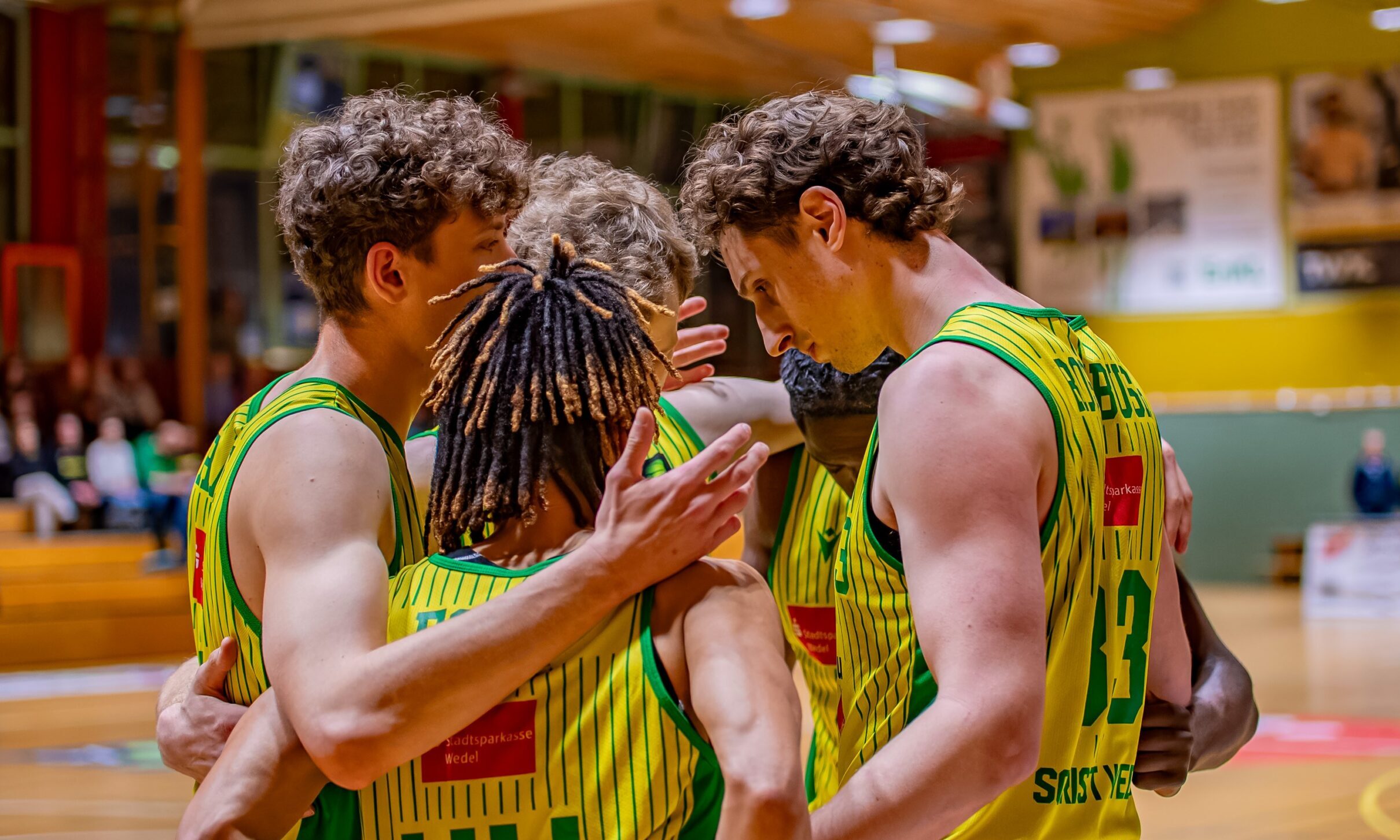 Vier Basketballspieler im gelb-grünen Trikot stehen im engen Kreis auf dem Spielfeld.