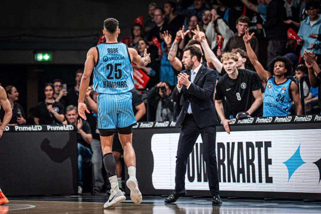 Basketballspieler im gestreiften Trikot mit der Rückennummer 25 läuft jubelnd an einem applaudierenden Mann im Anzug vorbei, im Hintergrund jubelnde Fans.