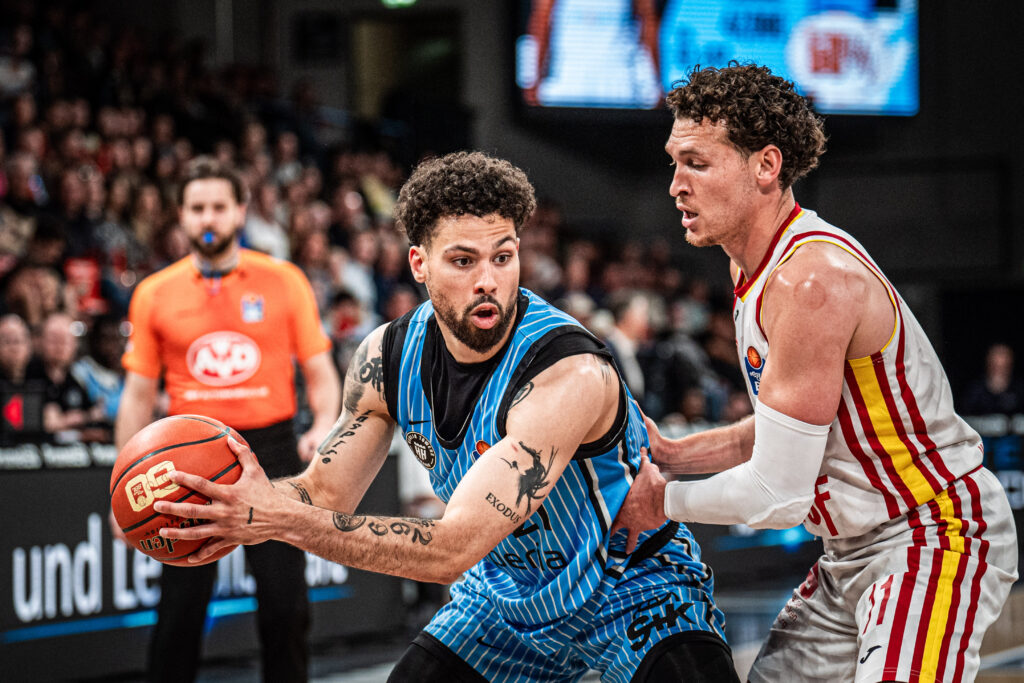 Basketballspieler im blauen Trikot hält den Ball und wird von einem Gegenspieler im weißen Trikot mit roten und gelben Streifen verteidigt.