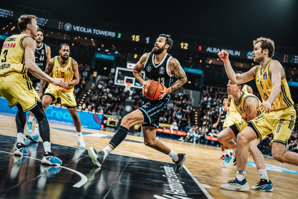 Basketballspieler in gelben und schwarzen Trikots verteidigen einen Spieler im schwarzen Trikot mit Ball auf dem Spielfeld vor Publikum.