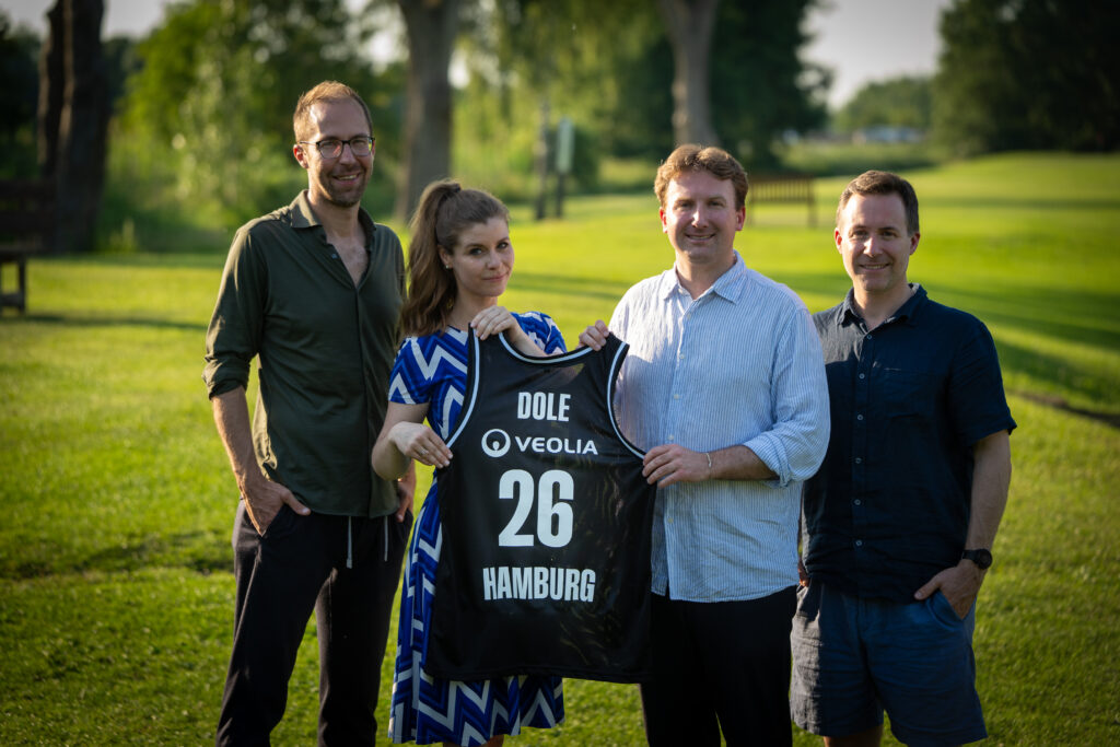 Vier Personen stehen auf einer Wiese, eine Person hält ein Basketballtrikot mit der Aufschrift 'DOLE', 'VEOLIA', '26' und 'HAMBURG' hoch.
